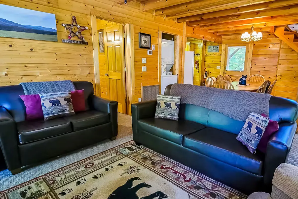 A warm living area view with black leather sofas, decorative pillows, and wooden walls, with the dining area visible in the background. The layout subtly connects the kitchen to communal spaces, suggesting proximity and easy socialization between cooking and gathering areas.