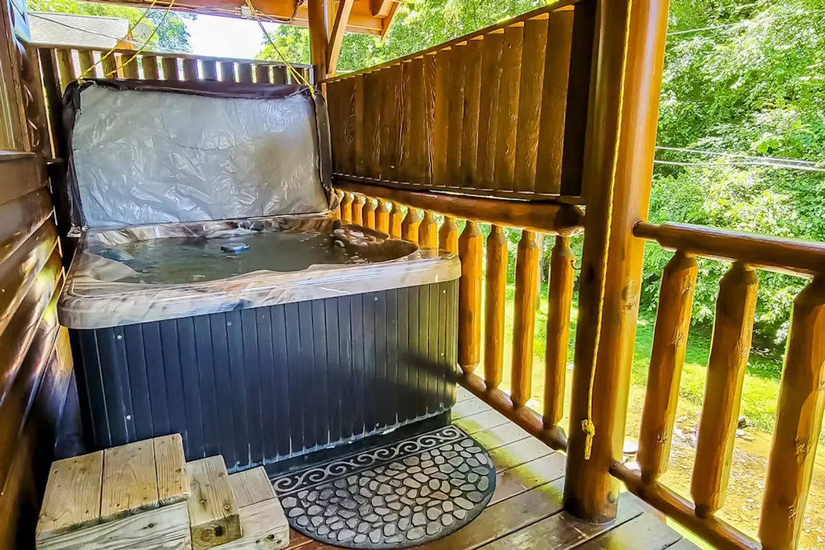 An inviting hot tub sits on a wooden balcony, overlooking the surrounding nature. The cozy setting features a stone mat and wooden railings, offering a private space to unwind and watch the sunset after a day of adventure.