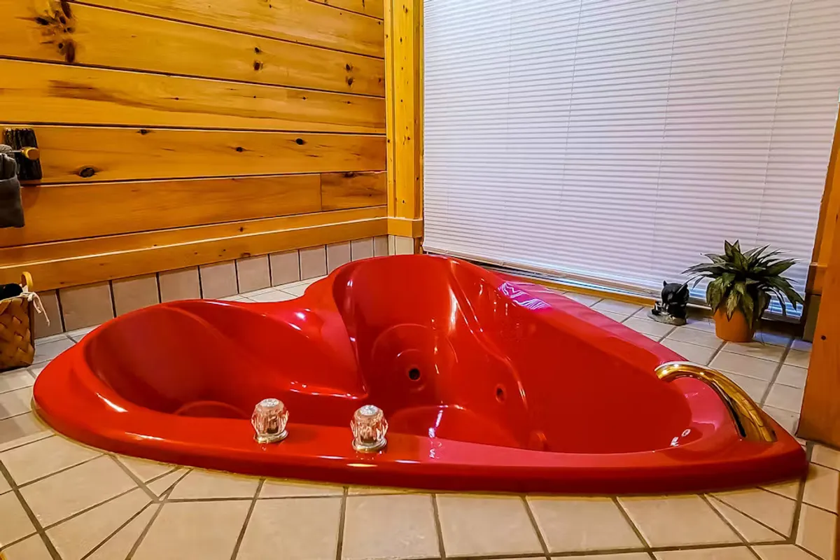 A cozy interior scene featuring a heart-shaped hot tub nestled against wooden walls with a decorative plant nearby. The lighting and ambiance set a romantic and intimate mood, ideal for couples seeking relaxation and connection.