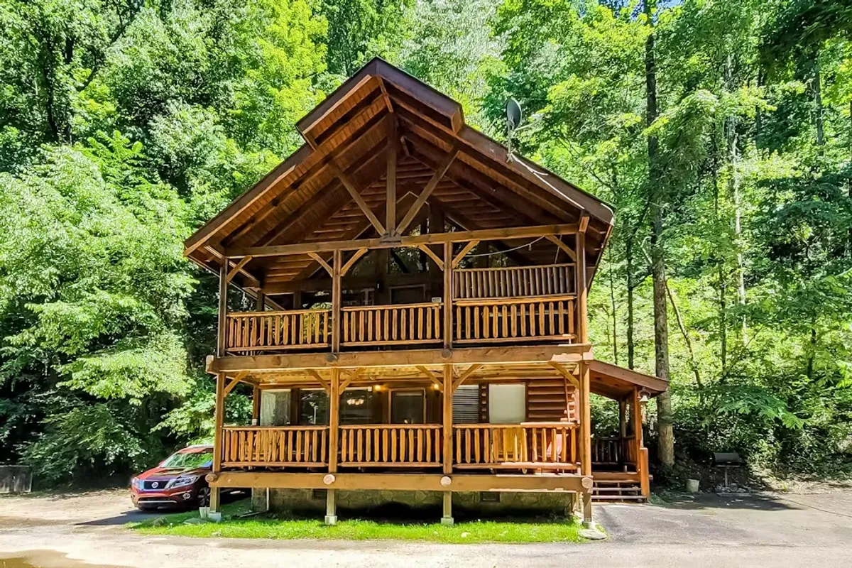 The hero image showcases a charming log cabin nestled amidst lush greenery, featuring a spacious wooden balcony and large windows that invite natural light. The serene setting is perfect for relaxation, with views of the surrounding forest and ample outdoor space for gathering, grilling, or simply unwinding with family and friends.