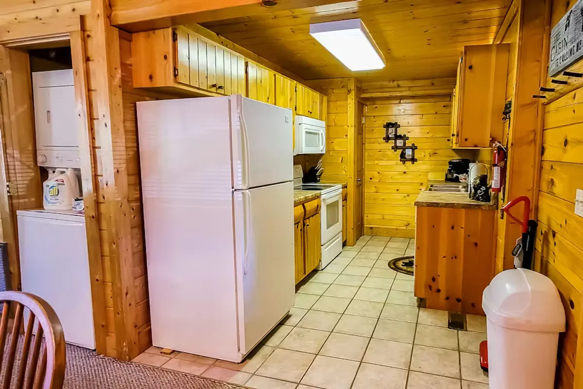 The image centers on a cozy and inviting kitchen space featuring a refrigerator, oven, wooden cabinetry, and a sink. The kitchen is well-lit and exudes warmth, offering ample space for meal preparation and gatherings. Modern appliances are visible, and the layout feels conducive to both quick snacks and more elaborate cooking activities.