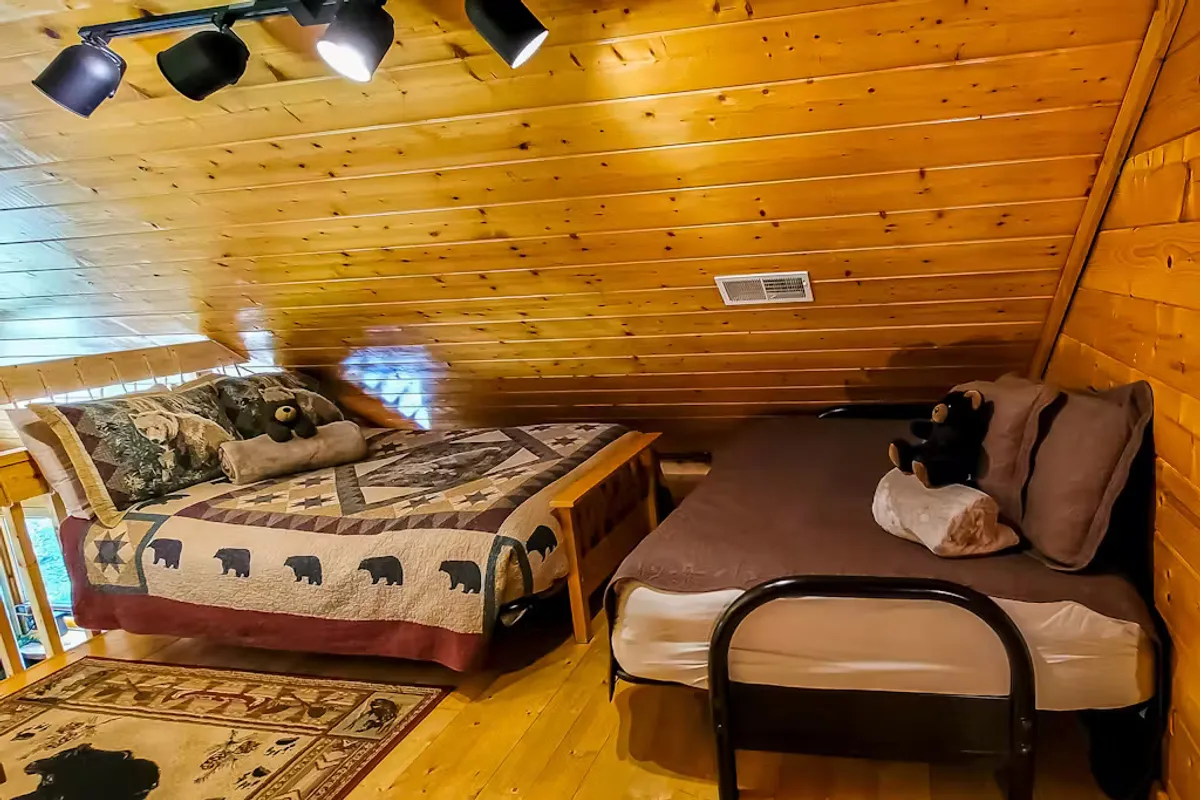 A welcoming bedroom with two beds adorned in decorative quilts, stuffed animals, and a rustic wooden ceiling. The space feels intimate and perfect for families or friends seeking comfort and togetherness.