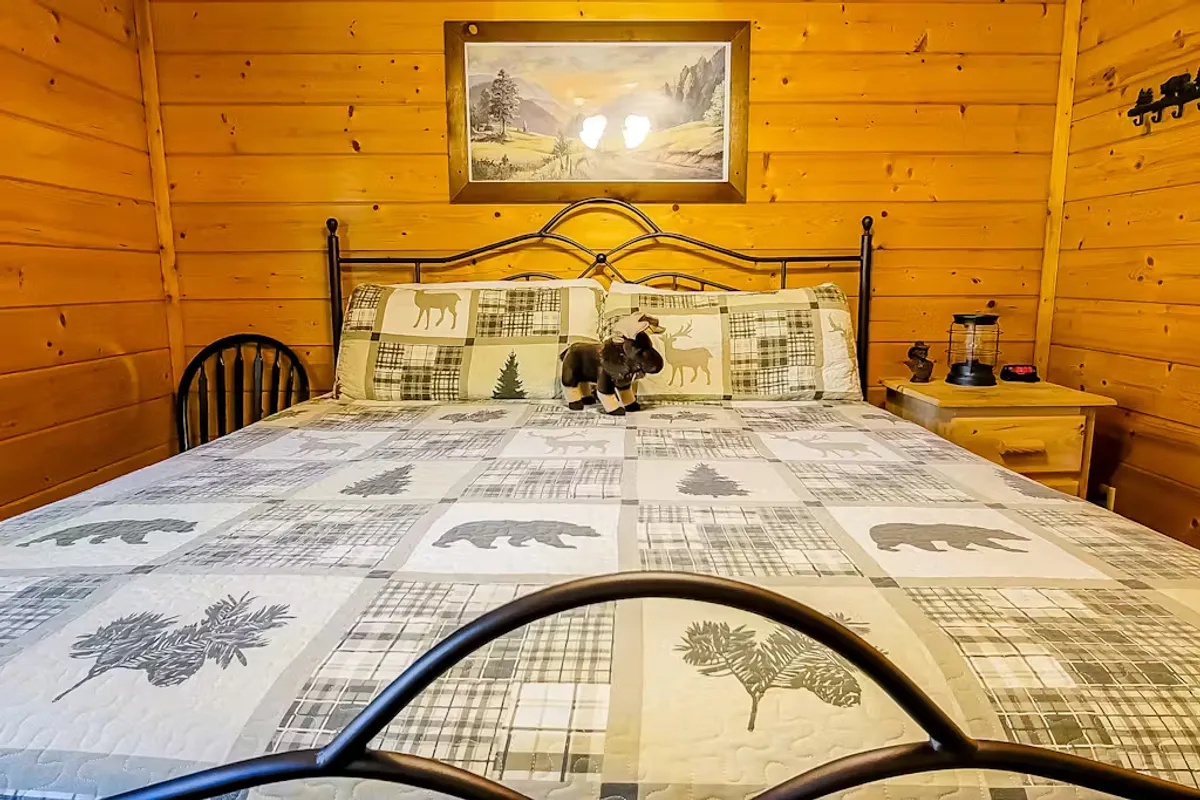 A warmly lit bedroom with a bed dressed in a patterned quilt, nature-themed artwork above the headboard, and a lantern on the nightstand, all surrounded by rustic wooden walls. The window offers a glimpse of greenery outside.
