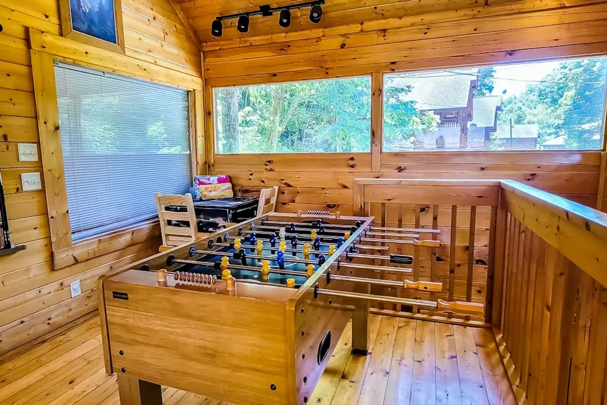 A lively corner of the game room featuring a foosball table, wood-paneled walls, comfortable chairs, and more large windows with views of surrounding greenery. The setup encourages playful interaction and socializing in a warm, rustic setting.